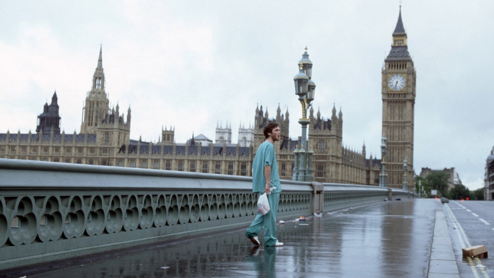 A man standing in a deserted London wearing hospital clothes, with the Big Ben in view behind him.
