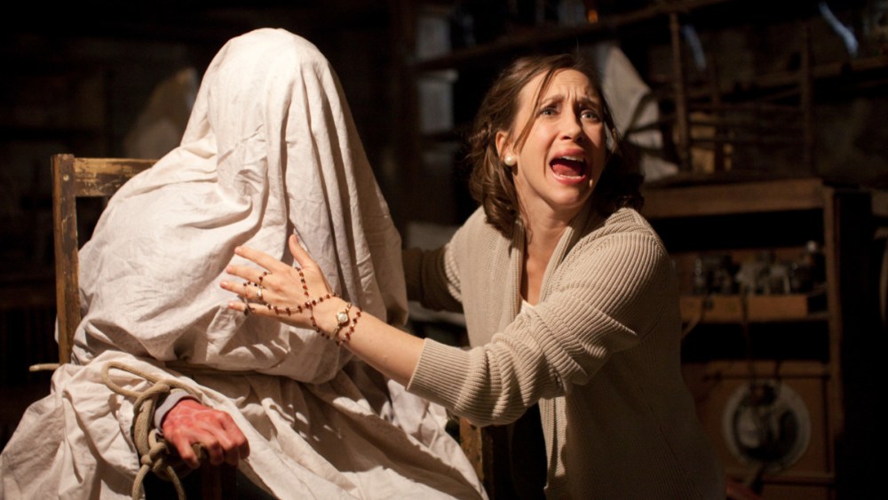 A person tied up to a chair with a blanket over them, while a woman besides them is screaming and trying to exorcise them.