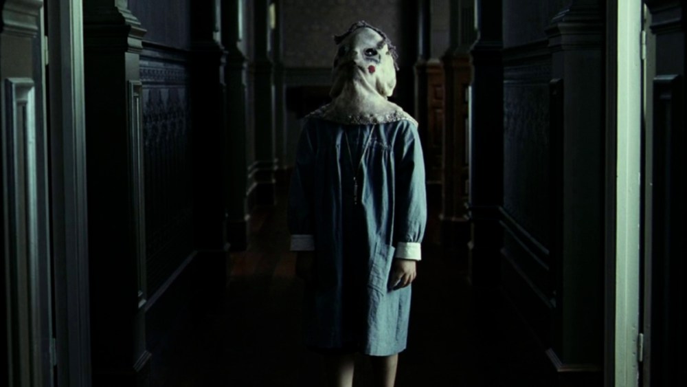 A young boy standing in a hallway with a nightgown and scary sheet over his head.